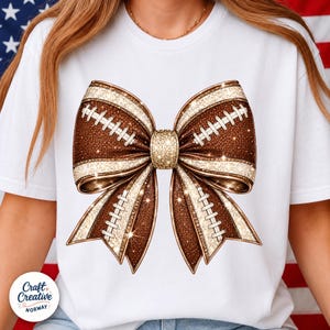 May include: A white t-shirt featuring a large graphic of a bow made from brown football-textured material with white laces and gold glitter accents. The center of the bow is a solid gold glitter knot, and the entire design has a sparkling finish.