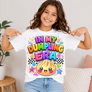 May include: A white short-sleeve t-shirt featuring a colorful graphic print with the text "IN MY DUMPLING ERA" in bold, bubble-style letters. Below the text is a cute, smiling dumpling illustration with pink cheeks, surrounded by stars.
