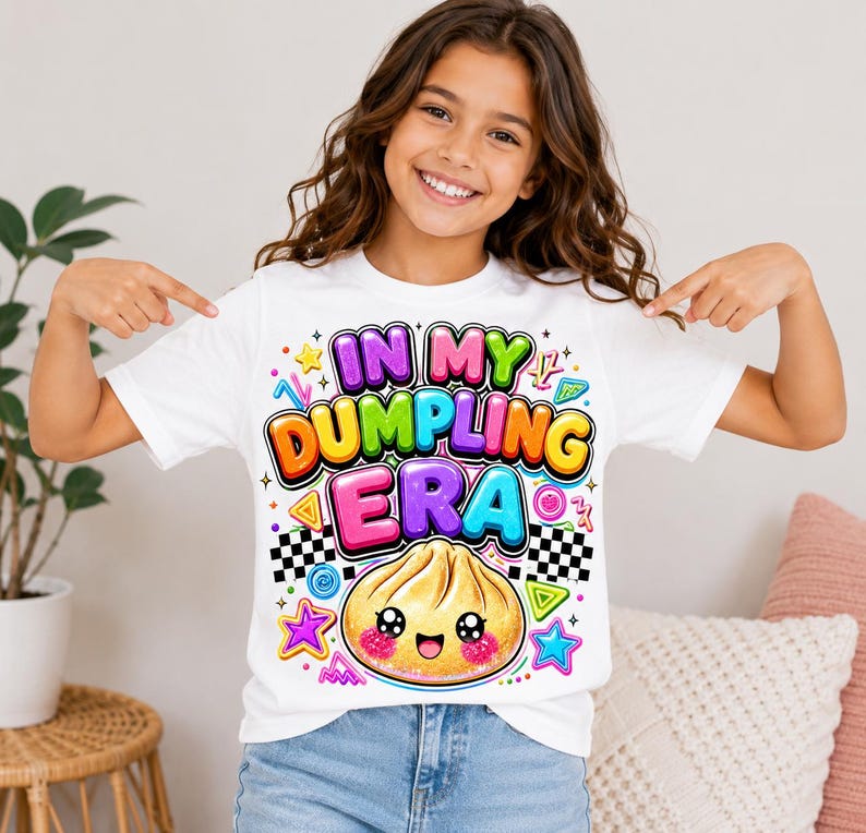 May include: A white short-sleeve t-shirt featuring a colorful graphic print with the text "IN MY DUMPLING ERA" in bold, bubble-style letters. Below the text is a cute, smiling dumpling illustration with pink cheeks, surrounded by stars.