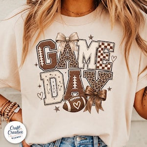 May include: A beige t-shirt featuring the text GAME DAY in large block letters. The letters incorporate football textures, leopard print, and checkered patterns. The design includes two bows, a football with a heart, and play diagrams.