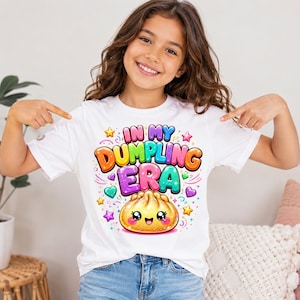 May include: A white short-sleeved t-shirt featuring a colorful graphic print that reads "IN MY DUMPLING ERA" in bold, bubble-style letters. Below the text is a cute, smiling dumpling character surrounded by small, vibrant stars and hearts.