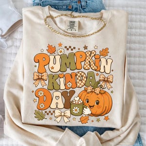 May include: A cream-colored crewneck sweatshirt featuring a graphic print with the text PUMPKIN KINDA DAY. The design includes a smiling orange pumpkin holding a mug, surrounded by bows, flowers, leaves, stars, and checkered patterns.