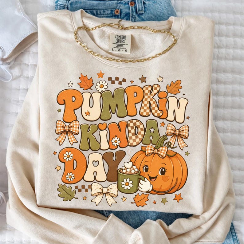 May include: A cream-colored crewneck sweatshirt featuring a graphic print with the text PUMPKIN KINDA DAY. The design includes a smiling orange pumpkin holding a mug, surrounded by bows, flowers, leaves, stars, and checkered patterns.