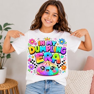 May include: A white short-sleeve t-shirt featuring a colorful graphic with the text "IN MY DUMPLING ERA" in bold, gradient lettering. The design includes a smiling, rainbow-colored dumpling character, small flowers, stars, and a black and white checkerboard pattern.