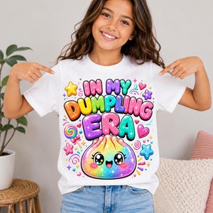 May include: A white short-sleeved t-shirt featuring a colorful graphic print with the text "IN MY DUMPLING ERA" in bubble letters. The design includes a smiling, rainbow-colored dumpling character, a lollipop, and various stars and hearts.