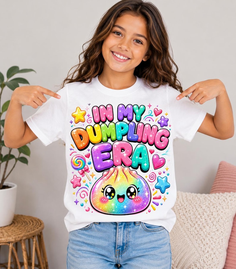 May include: A white short-sleeved t-shirt featuring a colorful graphic print with the text "IN MY DUMPLING ERA" in bubble letters. The design includes a smiling, rainbow-colored dumpling character, a lollipop, and various stars and hearts.