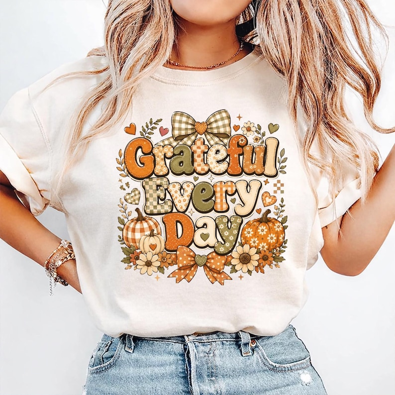 May include: A cream-colored t-shirt featuring a graphic print with the text "Grateful Every Day" in a retro font. The design includes orange and green checkered pumpkins, floral accents, and two plaid bows in a warm, autumnal color palette.