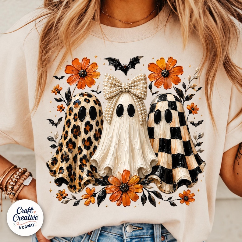 May include: A cream-colored t-shirt featuring a graphic of three ghosts. The left ghost has a leopard print pattern, the center ghost wears a large pearl-embellished bow, and the right ghost has a black and white checkerboard pattern. Orange flowers and small black bats surround the ghosts.