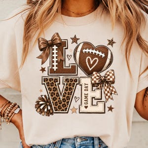May include: A beige t-shirt featuring a graphic print that spells LOVE. The L is topped with a brown bow, the O is a football heart, the V has a leopard print pattern, and the E includes the text GAME DAY with a checkered bow and pom-pom.