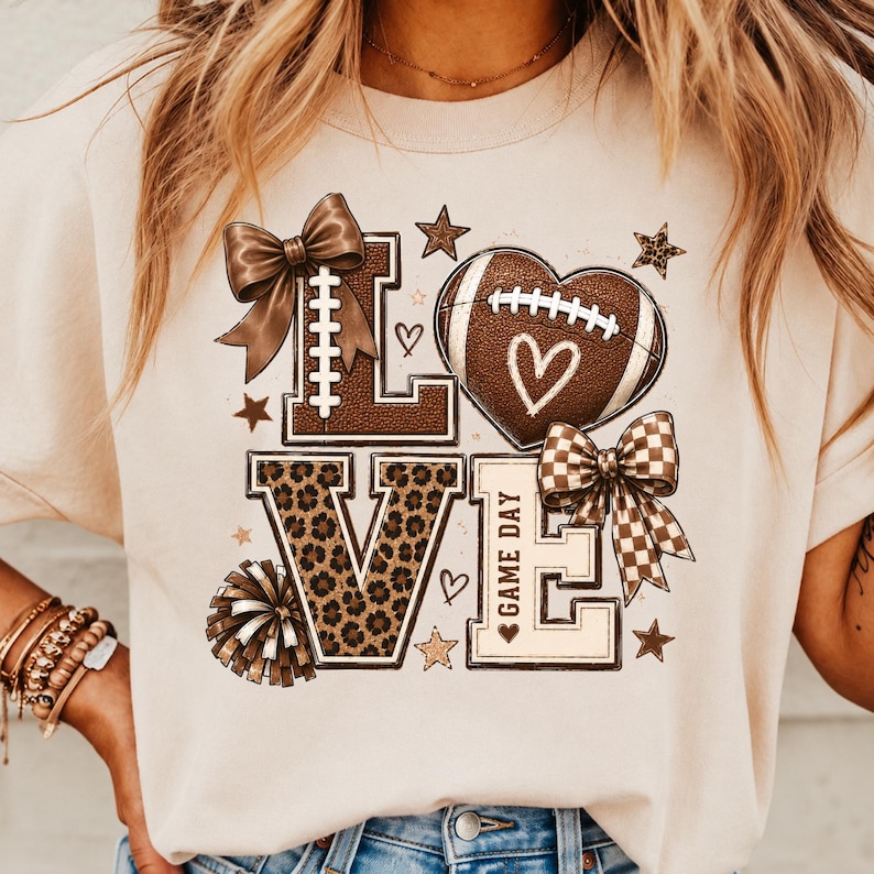 May include: A beige t-shirt featuring a graphic print that spells LOVE. The L is topped with a brown bow, the O is a football heart, the V has a leopard print pattern, and the E includes the text GAME DAY with a checkered bow and pom-pom.