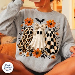 May include: A heather gray crewneck sweatshirt featuring a graphic of three ghosts. The left ghost has a leopard print, the center ghost wears a pearl-embellished bow, and the right ghost has a black and white checkerboard pattern, surrounded by orange flowers and bats.