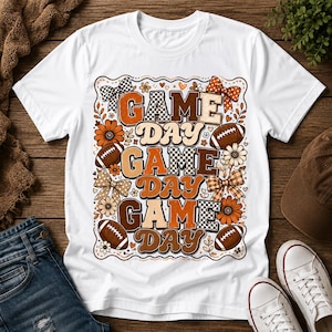 Op de afbeelding: Een wit t-shirt met korte mouwen en een grafische print met de herhaalde tekst "GAME DAY" in verschillende bruine en oranje lettertypes. Het ontwerp bevat illustraties van voetballen, ruitpatronen, bloemen en strikken in een rechthoek.