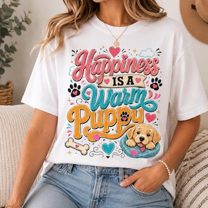 May include: A white t-shirt featuring a colorful graphic with the text "Happiness IS A Warm Puppy". The design includes a golden puppy resting in a blue bed, surrounded by pink and teal hearts, paw prints, and a bone illustration in a retro font style.