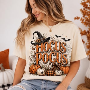 May include: A cream-colored short-sleeve t-shirt featuring a graphic print with the text HOCUS POCUS in orange letters. The design includes a black witch hat, pumpkins with polka dot and plaid patterns, a spider web, bats, and foliage accents.