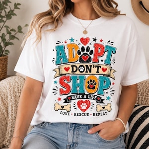 May include: A white short-sleeve t-shirt featuring a colorful graphic with the text ADOPT DON'T SHOP, SAVE A LIFE, and LOVE RESCUE REPEAT. The design includes paw prints, hearts, stars, and bone shapes in red, blue, orange, and black ink colors.