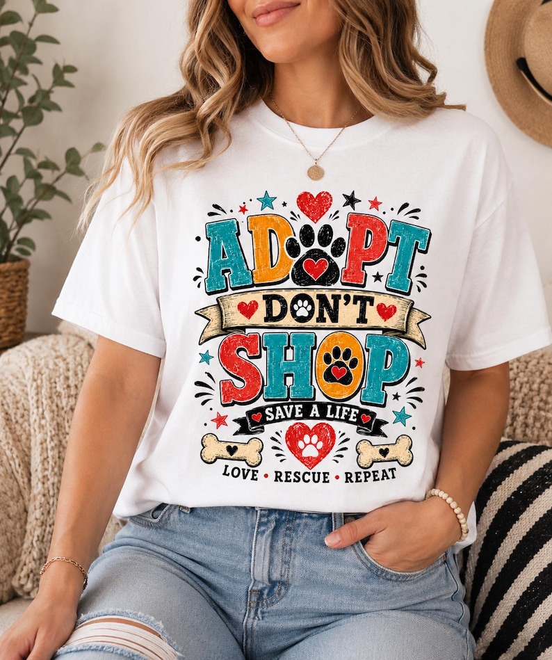 May include: A white short-sleeve t-shirt featuring a colorful graphic with the text ADOPT DON'T SHOP, SAVE A LIFE, and LOVE RESCUE REPEAT. The design includes paw prints, hearts, stars, and bone shapes in red, blue, orange, and black ink colors.