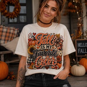 May include: A cream-colored short-sleeve t-shirt featuring a graphic print with the text "Fall Is My Favorite Color" in orange and black script. The design includes illustrations of pumpkins, sunflowers, autumn leaves, and a plaid bow.