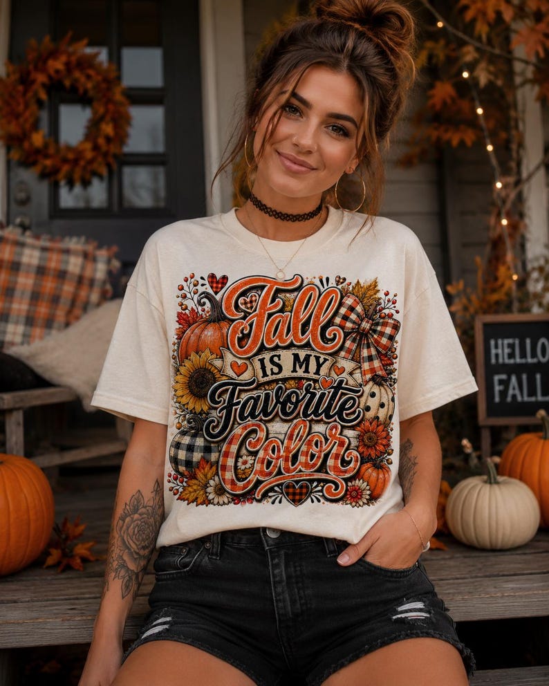 May include: A cream-colored short-sleeve t-shirt featuring a graphic print with the text "Fall Is My Favorite Color" in orange and black script. The design includes illustrations of pumpkins, sunflowers, autumn leaves, and a plaid bow.
