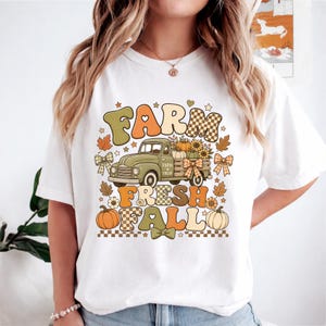 May include: A white t-shirt featuring a graphic print of a vintage green truck carrying pumpkins and sunflowers. The text "FARM FRESH FALL" is displayed in large, colorful, retro-style lettering, accented with small bows, leaves, and checkered patterns.