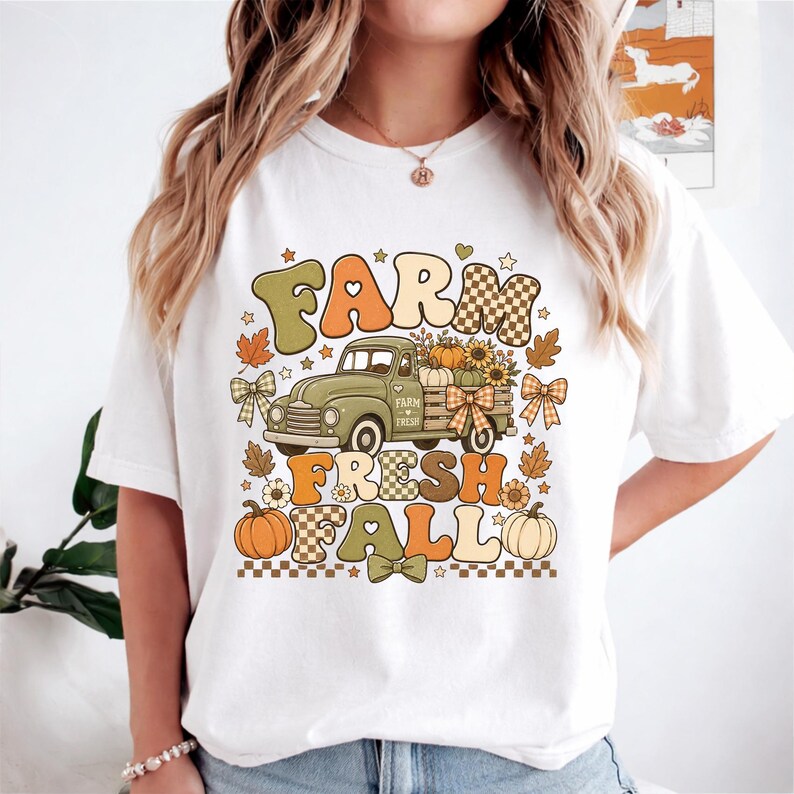 May include: A white t-shirt featuring a graphic print of a vintage green truck carrying pumpkins and sunflowers. The text "FARM FRESH FALL" is displayed in large, colorful, retro-style lettering, accented with small bows, leaves, and checkered patterns.