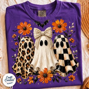 May include: A purple t-shirt featuring a graphic of three ghosts. The left ghost has a leopard print, the center ghost wears a large pearl-covered bow, and the right ghost has a black and white checkerboard pattern. Orange flowers and bats surround them.