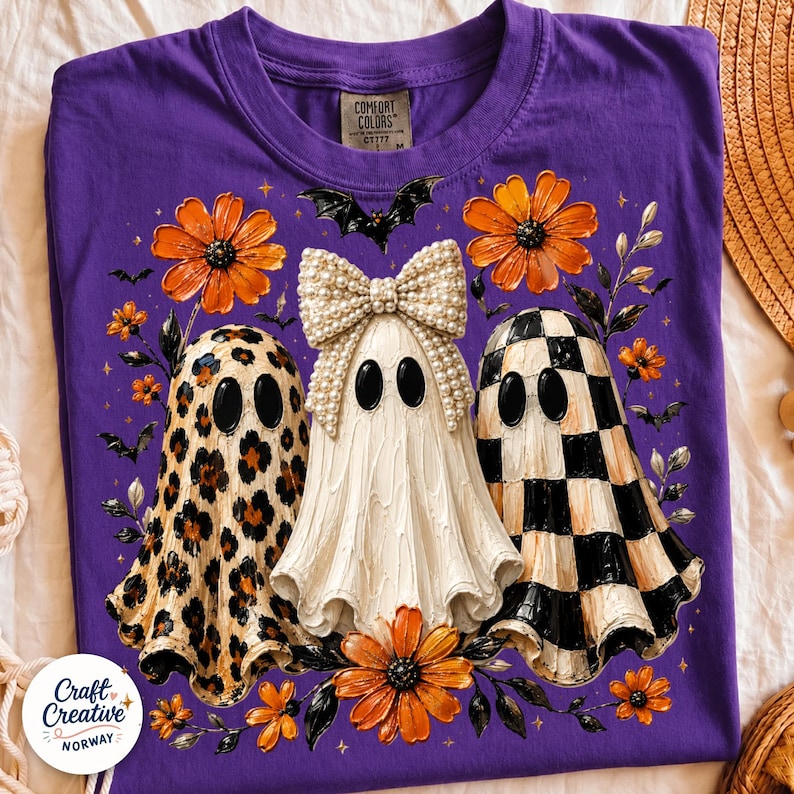 May include: A purple t-shirt featuring a graphic of three ghosts. The left ghost has a leopard print, the center ghost wears a large pearl-covered bow, and the right ghost has a black and white checkerboard pattern. Orange flowers and bats surround them.