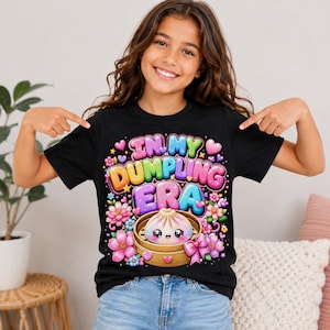 May include: A black t-shirt featuring a colorful graphic with the text "IN MY DUMPLING ERA" in bubble letters. The design includes a cute, smiling dumpling in a bamboo steamer, surrounded by pink flowers, small hearts, and stars in vibrant colors.