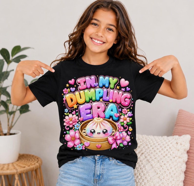 May include: A black t-shirt featuring a colorful graphic with the text "IN MY DUMPLING ERA" in bubble letters. The design includes a cute, smiling dumpling in a bamboo steamer, surrounded by pink flowers, small hearts, and stars in vibrant colors.
