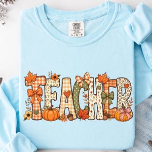 May include: A light blue long-sleeve t-shirt featuring the word TEACHER in large, decorative letters. Each letter is filled with autumn-themed patterns including orange plaid, leopard print, pumpkins, maple leaves, bows, and small flowers.