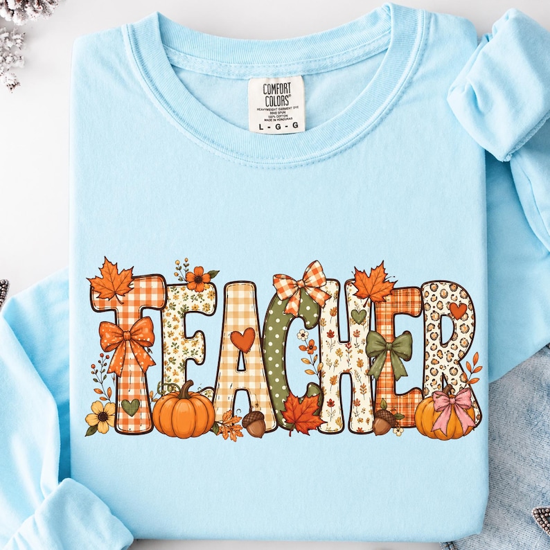 May include: A light blue long-sleeve t-shirt featuring the word TEACHER in large, decorative letters. Each letter is filled with autumn-themed patterns including orange plaid, leopard print, pumpkins, maple leaves, bows, and small flowers.