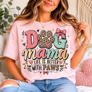 May include: A light pink t-shirt with the words "Dog Mama" in a colorful, playful design. The design includes a paw print, hearts, and the phrase "Life is Better With Paws".