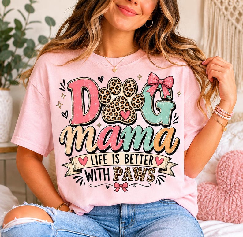 May include: A light pink t-shirt with the words "Dog Mama" in a colorful, playful design. The design includes a paw print, hearts, and the phrase "Life is Better With Paws".