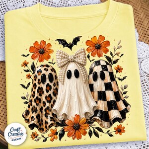 May include: A pale yellow t-shirt featuring a graphic of three ghosts. One ghost has a leopard print, one wears a large pearl-embellished bow, and one has a black and white checkerboard pattern. The design includes orange flowers and bats.