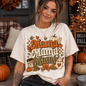 May include: A cream-colored short-sleeve t-shirt featuring a graphic print with the repeated word Mama in orange, cream, and olive green. The design includes illustrations of pumpkins, autumn leaves, a coffee cup, and a mug with whipped cream.