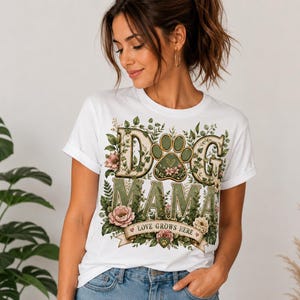 May include: White t-shirt with a floral design and the words "DOG MAMA" in green and beige. The design includes a dog paw print and the text "LOVE GROWS HERE" on a banner. The shirt has rolled-up sleeves.