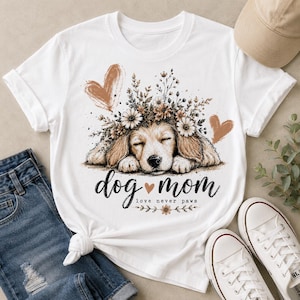May include: White t-shirt featuring a graphic of a sleeping dog with a floral crown and two hearts. The text "dog mom love never paws" is below the dog illustration. The shirt is paired with blue jeans and white sneakers.