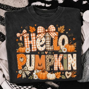 May include: A black t-shirt featuring the text Hello Pumpkin in large, decorative letters. The letters are filled with orange and white gingham, leopard print, and floral patterns. The design includes orange pumpkins, bows, and autumn leaves.