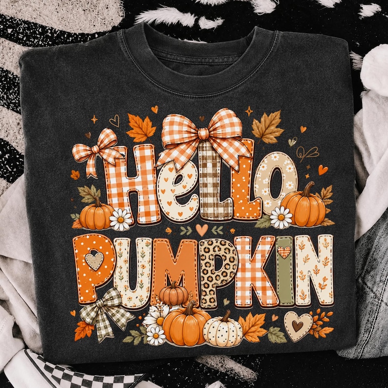 May include: A black t-shirt featuring the text Hello Pumpkin in large, decorative letters. The letters are filled with orange and white gingham, leopard print, and floral patterns. The design includes orange pumpkins, bows, and autumn leaves.
