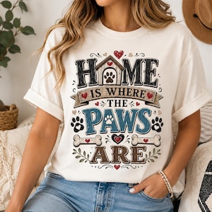 May include: A cream-colored short-sleeve t-shirt featuring a graphic design with the text "HOME IS WHERE THE PAWS ARE". The design includes a dog house, paw prints, bones, and red hearts, with the word "HOME" in black, "PAWS" in blue, and "ARE" in brown.