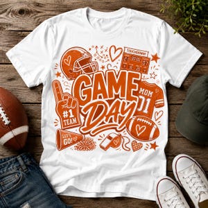 Op de afbeelding: Een wit t-shirt met korte mouwen en een grote oranje afbeelding met de tekst "GAME DAY". Het ontwerp bevat illustraties van een footballhelm, een scorebord, een schuimvinger, een shirt met "MOM", een football, een megafoon en een fluitje.