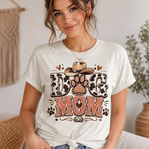 May include: A light beige t-shirt featuring the text "DOG MOM" in a Western-style design. The design includes a cowboy hat, paw prints, and hearts. The shirt is worn by a person with their hands in their pockets.