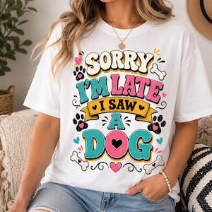 May include: A white short-sleeve t-shirt featuring a colorful graphic print that reads "SORRY I'M LATE I SAW A DOG" in bold, retro-style lettering. The design includes paw prints, bones, and small hearts in shades of pink, teal, and yellow.