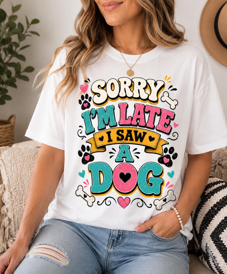 May include: A white short-sleeve t-shirt featuring a colorful graphic print that reads "SORRY I'M LATE I SAW A DOG" in bold, retro-style lettering. The design includes paw prints, bones, and small hearts in shades of pink, teal, and yellow.