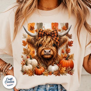 Può includere: Una t-shirt beige con la grafica di una mucca highland pelosa con un fiocco a quadri tra le corna. La mucca è circondata da zucche arancioni e bianche e foglie autunnali su uno sfondo a strisce verticali nei toni del marrone e dell'arancione.