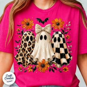 May include: A bright pink t-shirt featuring a graphic of three ghosts. The left ghost has a leopard print pattern, the center ghost is cream-colored with a large pearl-embellished bow, and the right ghost has a black and white checkerboard pattern.