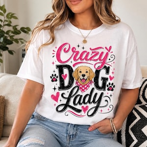 May include: A white short-sleeved t-shirt featuring a graphic print with the text "Crazy Dog Lady" in black and pink lettering. The design includes a golden retriever illustration wearing a pink bandana, surrounded by pink hearts and black paw prints.
