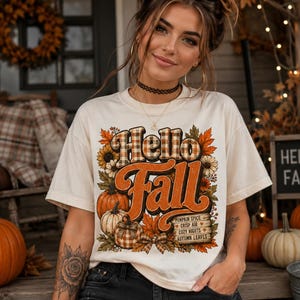 May include: A cream-colored short-sleeve t-shirt featuring a graphic print with the text Hello Fall in orange and plaid lettering. The design includes illustrations of pumpkins, sunflowers, autumn leaves, and a sign listing Pumpkin Spice, Crisp Air, Cozy Nights, and Autumn Leaves.