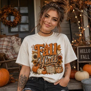 May include: A cream-colored short-sleeve t-shirt featuring a graphic print with the text Fall Y'all, surrounded by illustrations of sunflowers, pumpkins, and autumn leaves, along with a small sign listing Sweater Weather, Pumpkin Kisses, and Autumn Wishes.