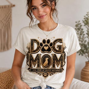 May include: Cream-colored t-shirt with the text "DOG MOM" in a decorative gold and black design. The design includes a paw print, a heart, and a silhouette of a dog. The text "LOYAL. LOVED. MY GREATEST MASTERPIECE" is also included.