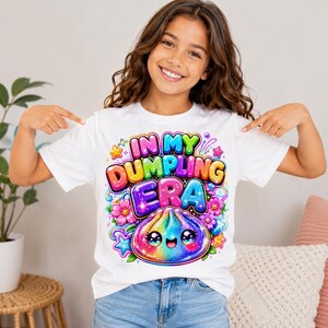 May include: A white short-sleeved t-shirt featuring a colorful graphic print with the text "IN MY DUMPLING ERA" in bold, bubble-style letters. The design includes a smiling, rainbow-colored dumpling character surrounded by pink flowers and stars.